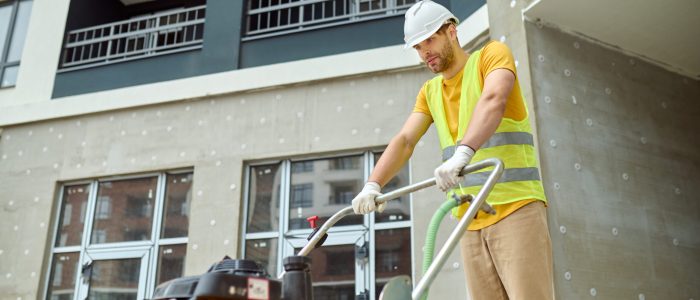 Effort, fatigue. Young adult tired man in protective helmet and bright vest with special equipment working on construction site against background of new building